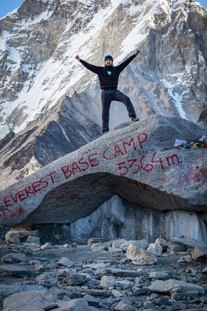 Traveler posing at Everest Base Camp with Mount Everest visible in the distance.